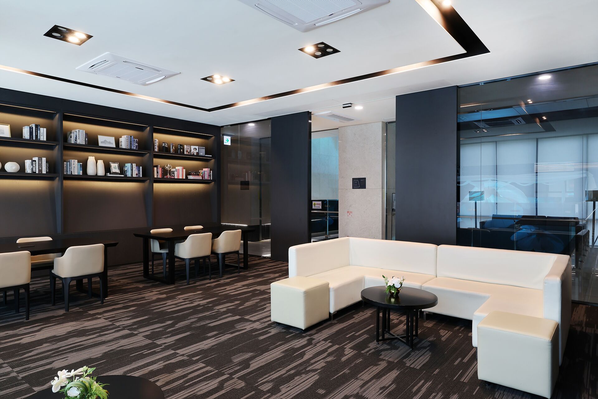 A modern lounge area with a sofa, chairs, a bookshelf, and a glass wall.