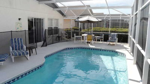 A covered outdoor swimming pool with lounge chairs and umbrellas, surrounded by a white fence. - Hampton Lakes Homes (Davenport)