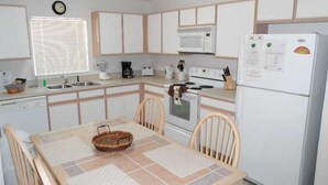 A compact kitchen with white cabinets, a microwave, a stove, and a refrigerator. There is a dining table with chairs and a basket on the table. - Hampton Lakes Homes (Davenport)