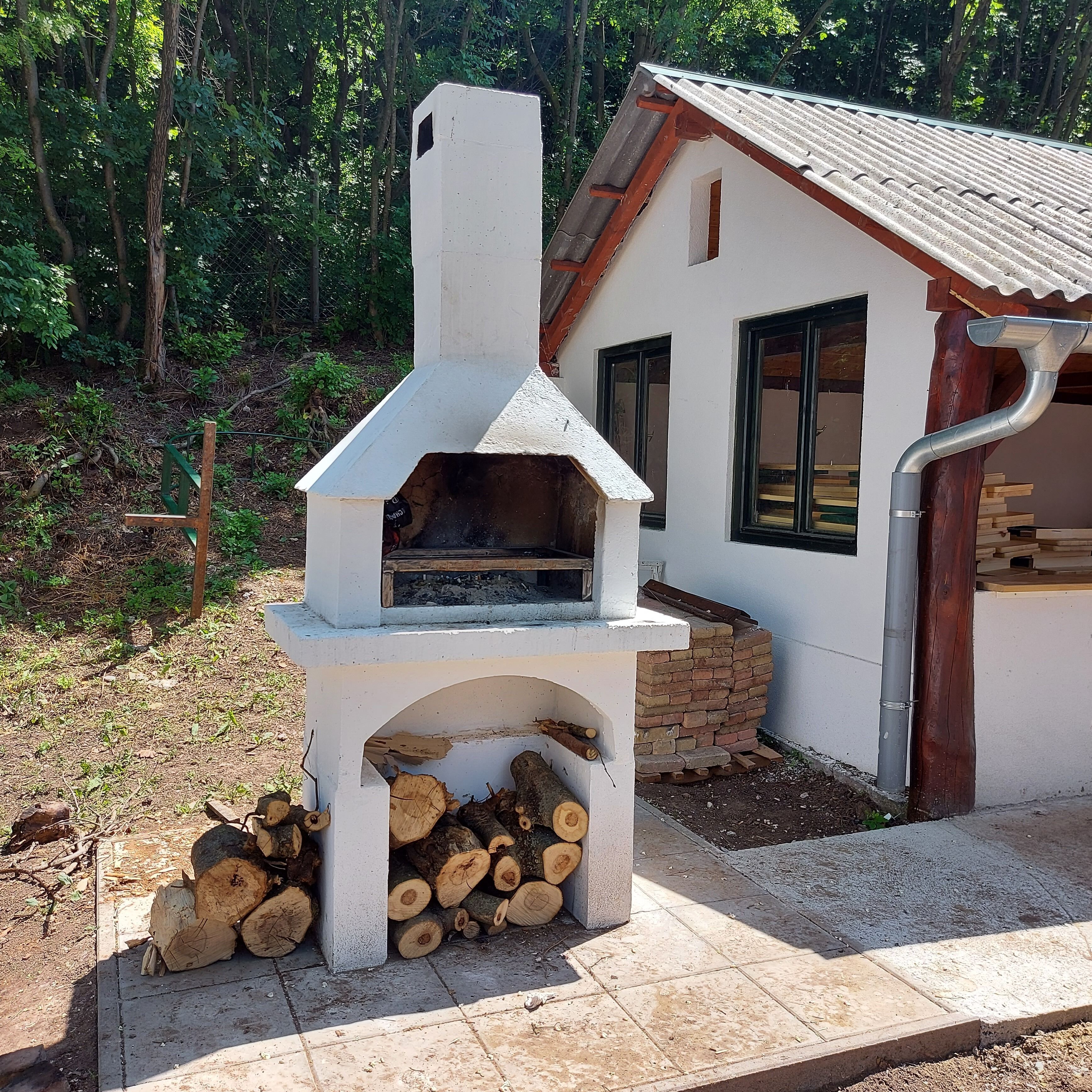 Eine rustikale Außenküche mit Holzofen, Holzstapel und einem Haus im Hintergrund.