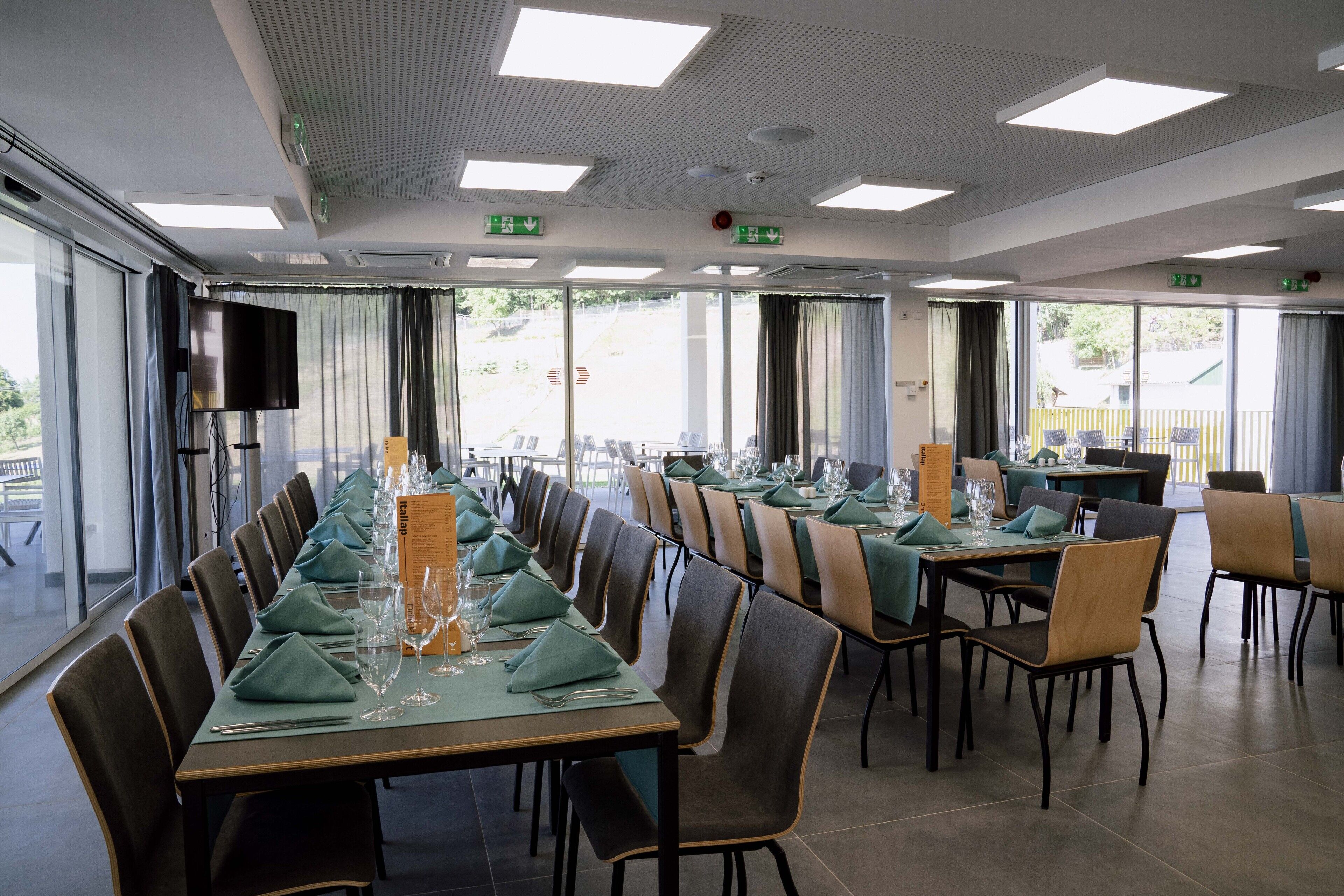 Uma sala de jantar com mesas compridas preparadas para uma refeição, equipadas com cadeiras, toalhas de mesa e talheres.