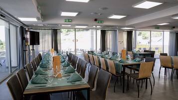 Uma sala de jantar com mesas compridas preparadas para uma refeição, equipadas com cadeiras, toalhas de mesa e talheres.