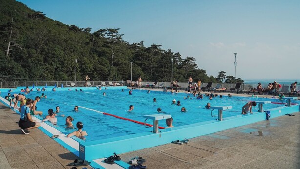 Ein Schwimmbecken mit mehreren Bahnen, umgeben von einer gefliesten Liegefläche und einem bewaldeten Hügelhang.