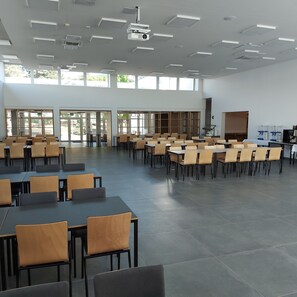 A spacious conference room with multiple tables and chairs, large windows, and ceiling-mounted projectors.