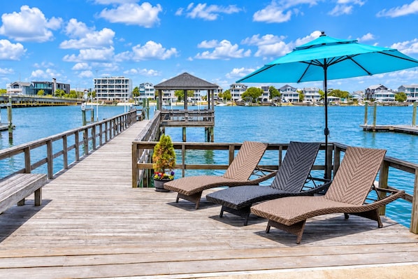 A wooden pier with lounge chairs and a gazebo, overlooking a body of water with buildings in the background.