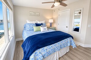 A bedroom with a bed, a nightstand, a lamp, a window with a view, a ceiling fan, and a door. - Experience the island lifestyle, live like a local! 4Br/3.5ba waterfront duplex. (Wrightsville Beach)