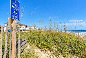 Una playa con un cartel de acceso público, una valla de madera y casas al fondo.
