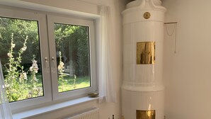 Una habitación con un ventanal grande, un radiador blanco, una silla gris, un objeto cilíndrico blanco con detalles dorados y una cesta de mimbre con objetos.