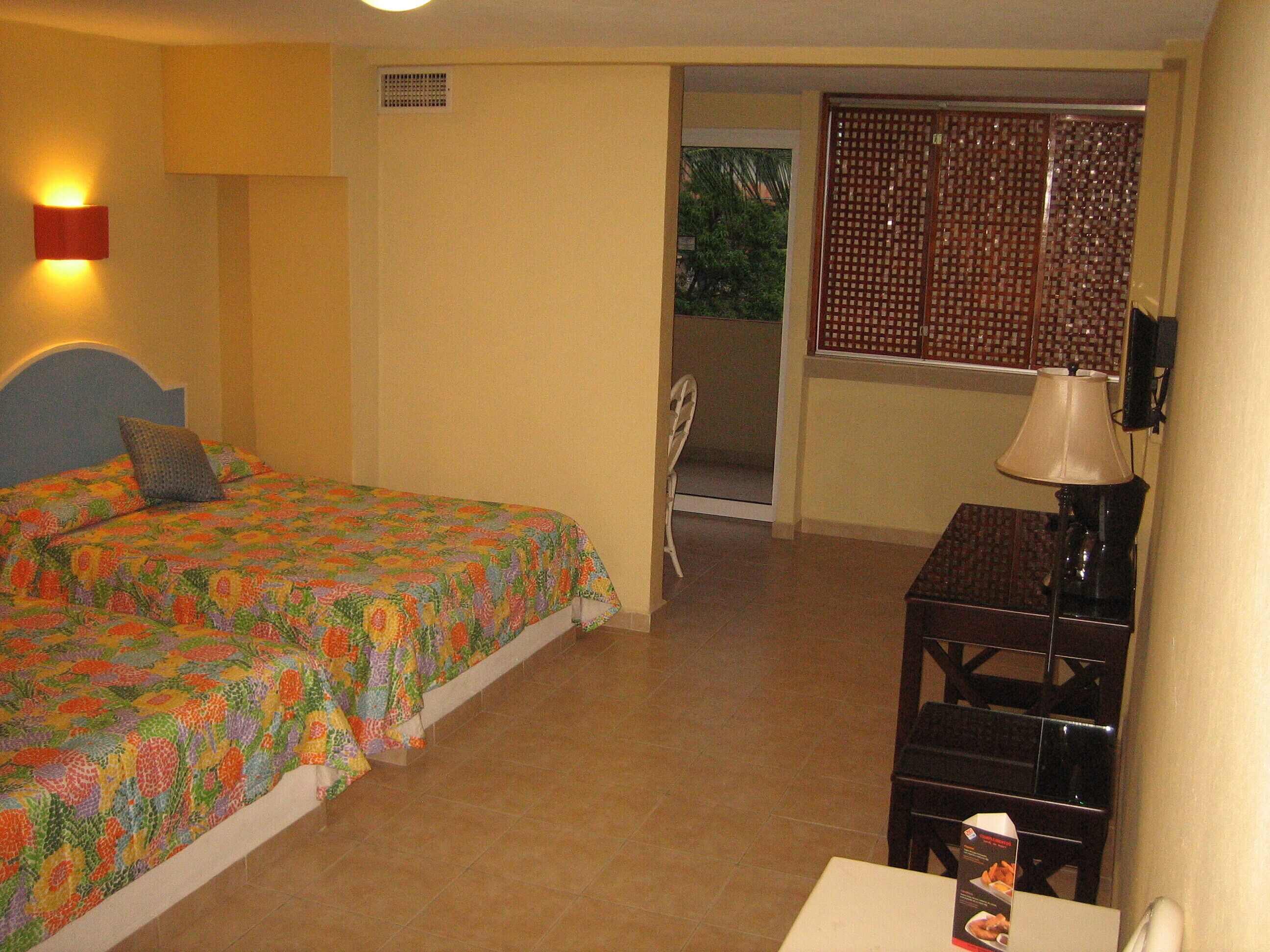 A hotel room with a bed, a desk, a chair, a lamp, and a window with a view of greenery outside.