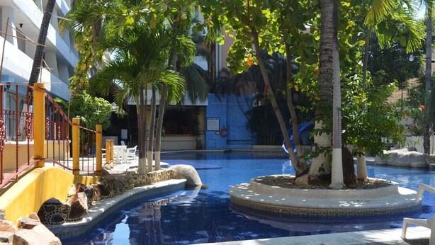 Zona de piscina con fuente circular, palmeras y barandilla amarilla.