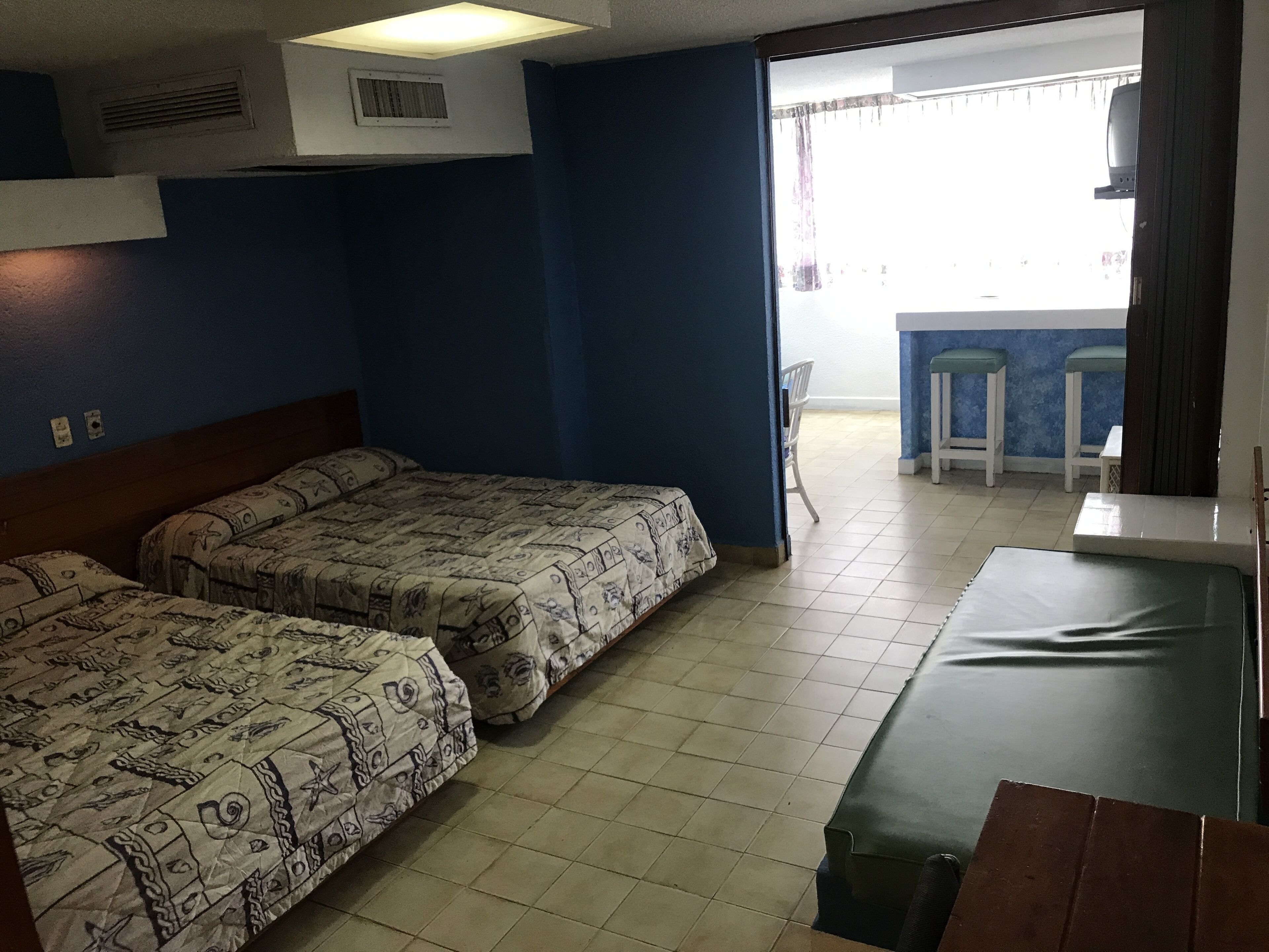 Una habitación con dos camas, una mesita pequeña y una silla. Hay una ventana con cortinas, una rejilla de ventilación en el techo y una puerta que da a otra habitación.