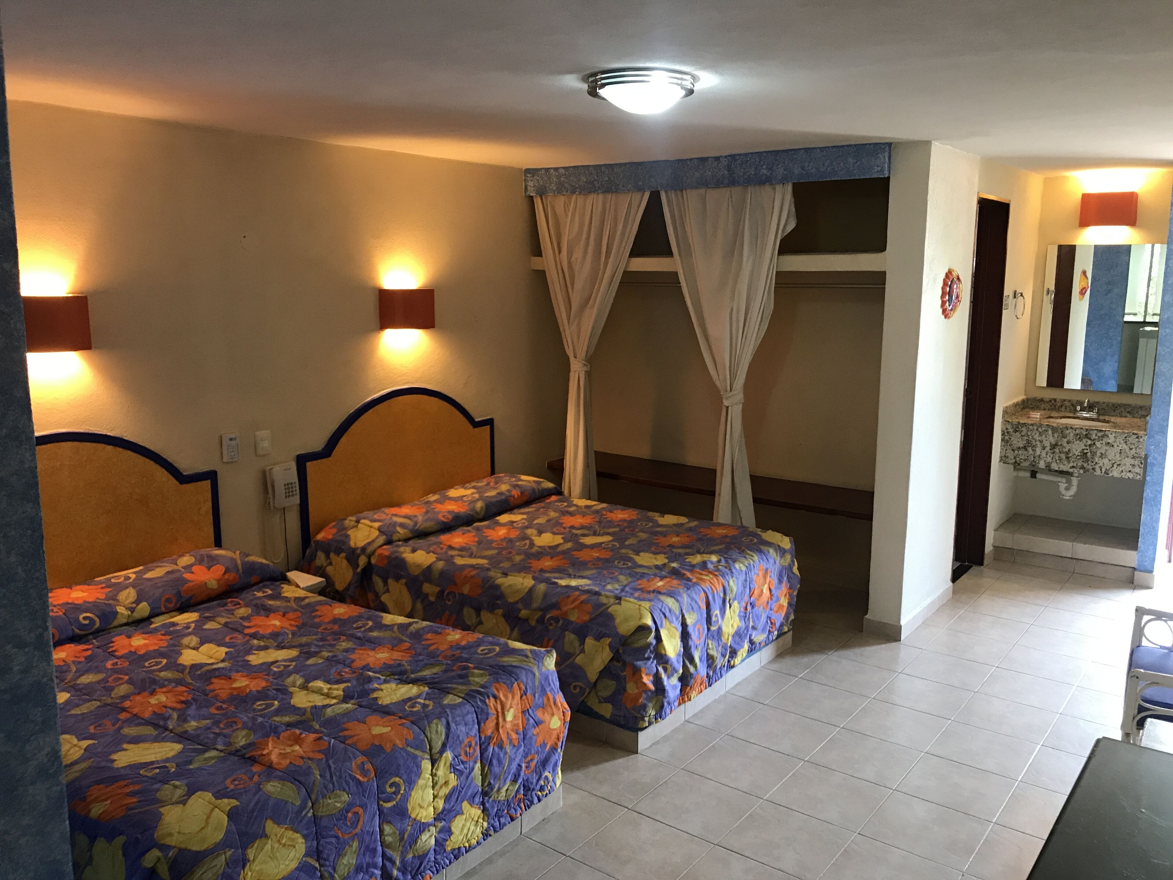 Quarto de hotel com duas camas, banheiro com pia e espelho, e uma luminária no teto.