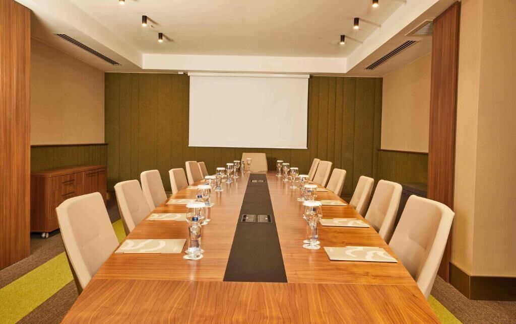Un salón de conferencias con una larga mesa, sillas, una pantalla de proyector y un atril.
