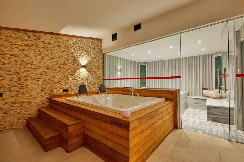 A modern bathroom with a large wooden bathtub, a stone wall, and a glass shower enclosure.