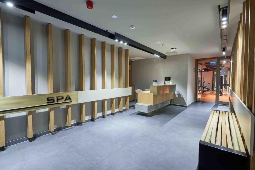 A reception area with a wooden reception desk labeled 'SPA', a seating area, and a corridor leading to other parts of the hotel.