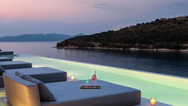An infinity pool with a modern lounge area, a candle, and a view of the water and hills.