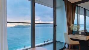 A modern hotel room with a large balcony, a round table, a chair, a desk with a phone, and a view of the sea and distant mountains. - Grand Gosia Hotel (Nha Trang)