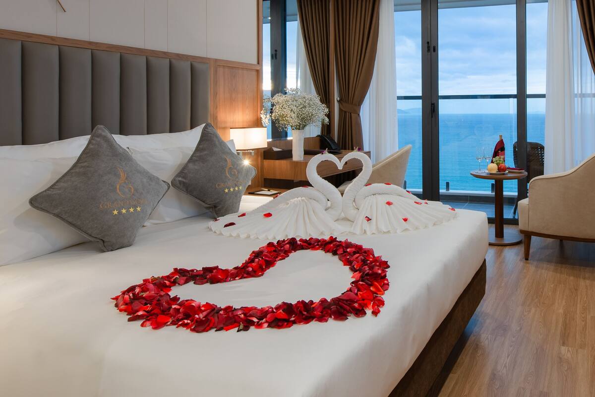 Hotelkamer met een groot bed, een hartvormige bloemblaadjesdecoratie van rode rozen, een boeket witte bloemen en uitzicht op de oceaan door grote ramen.