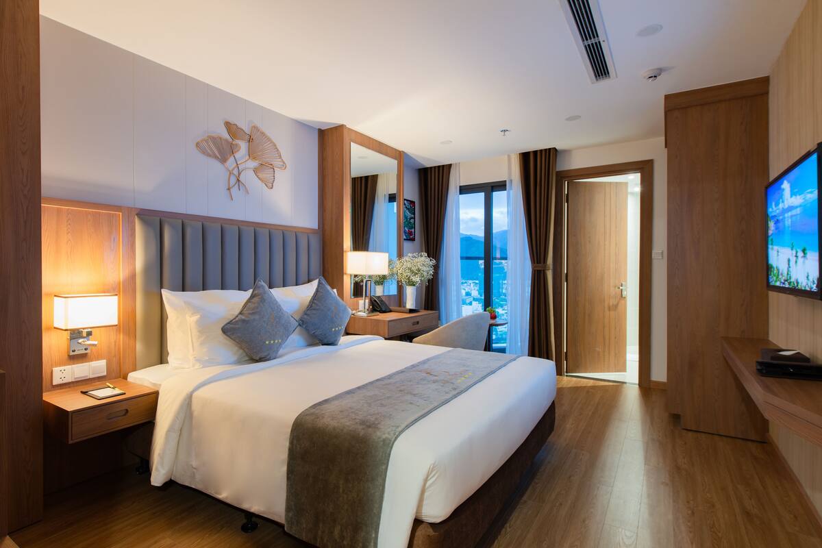 Een hotelkamer met een groot bed, een nachtkastje met een lamp, een televisie en uitzicht op het buiten.
