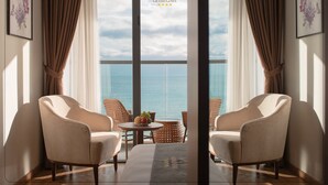 A room with two beige armchairs facing a large window overlooking the sea, a small round table with fruit, and wicker chairs. - Grand Gosia Hotel (Nha Trang)