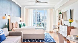 A modern hotel room with a large bed, a desk, a sofa, and a view of the city skyline through large windows. - Blue Accent Studio Suite | Bay & City Views (Miami)
