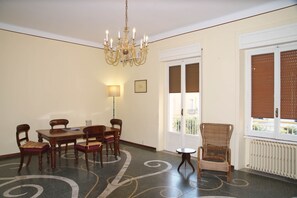A dining area with a table and chairs, a chandelier, a window with blinds, and a radiator. - 2120 Appartamento Roberto (Gallipoli)