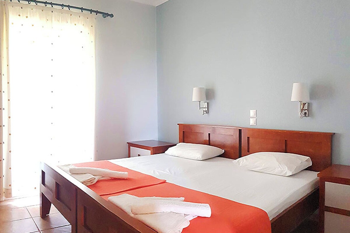 A neatly made bed with white linens and a red bedspread, two bedside tables with lamps, and a window with curtains.
