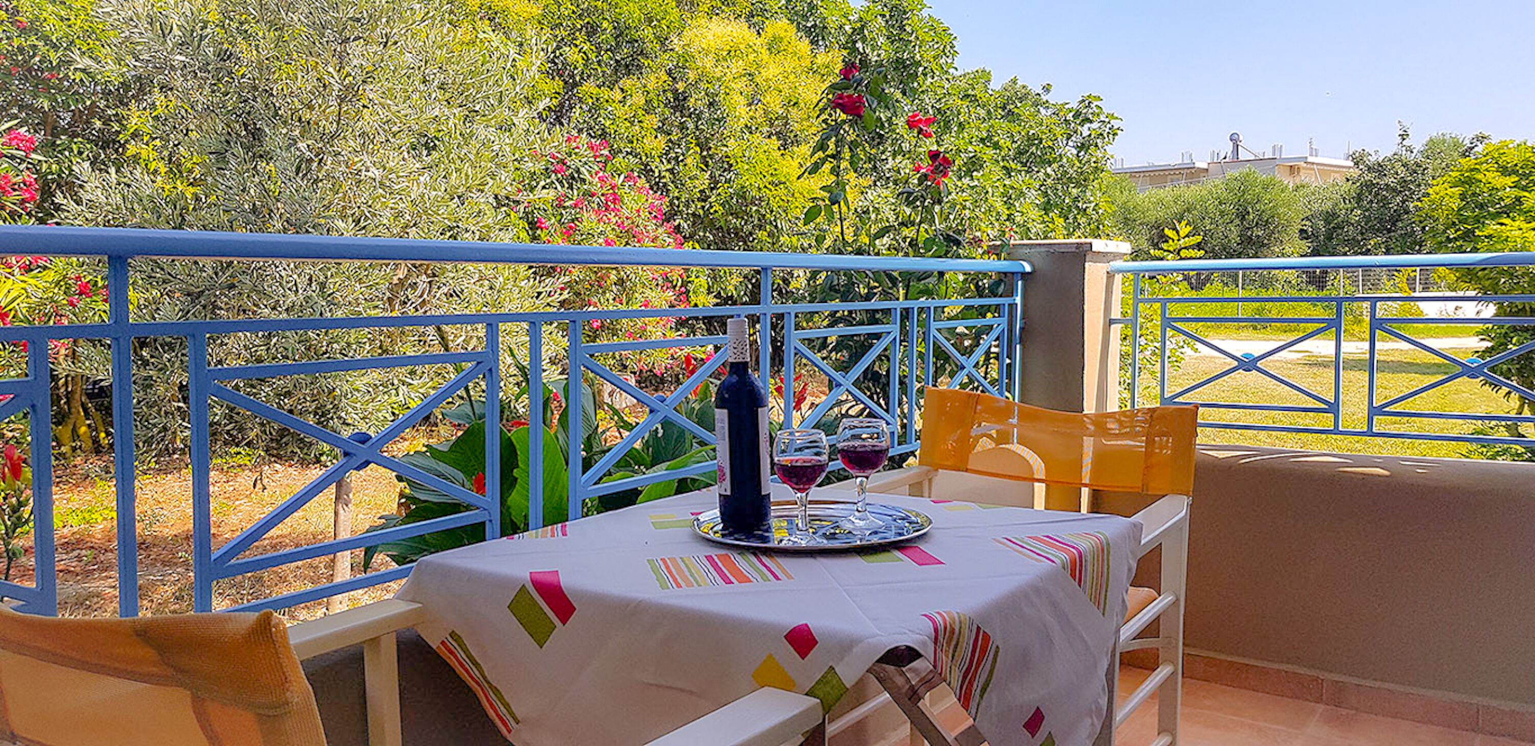 A table set for two with a bottle and glasses, blue metal railings, and a view of greenery and buildings in the background.