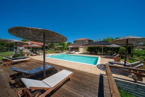 A wooden deck with lounge chairs and umbrellas, a swimming pool, and a house in the background. - Aggelina's Apartments (Kefalonia)