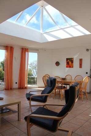 Une pièce spacieuse avec un plafond haut et une lucarne, comprenant une table à manger avec des chaises, un canapé et une vue sur l’extérieur.