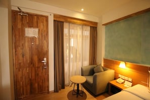 A hotel room with a wooden door, a small round table, a sofa, a bed, and a nightstand with a lamp. - Liberta Hotel Jimbaran (Kedonganan)