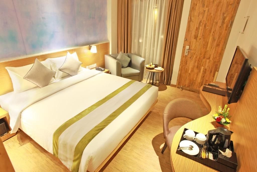 Habitación de hotel con cama, escritorio con cafetera, silla, pequeña mesa con un arreglo floral y televisor en la pared.