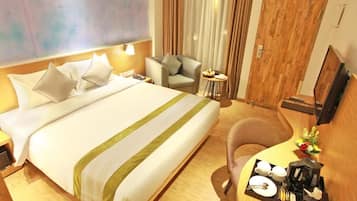 Habitación de hotel con cama, escritorio con cafetera, silla, pequeña mesa con un arreglo floral y televisor en la pared.