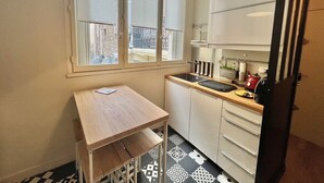 A compact kitchen with a small table and cabinets, a window with blinds, and a hanging light fixture. - Jaures 2 bedrooms 7 minuts walking from Palais (Cannes)