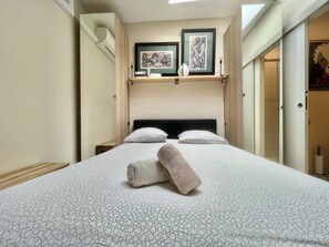 A hotel room with a bed, two pillows, a bedside table, and a wall-mounted shelf with framed artwork.