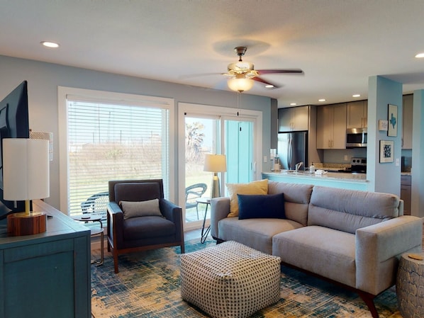 A modern living room with a sofa, armchair, and ottoman, a dining area with a table and chairs, and a kitchen with cabinets and appliances.