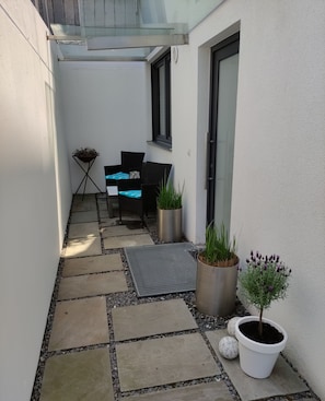 A narrow outdoor patio with a seating area, potted plants, and a small table.