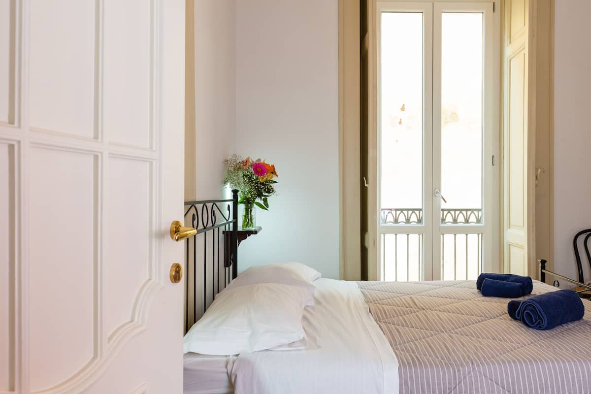 A hotel room with a bed, a nightstand with a vase of flowers, and a balcony door.