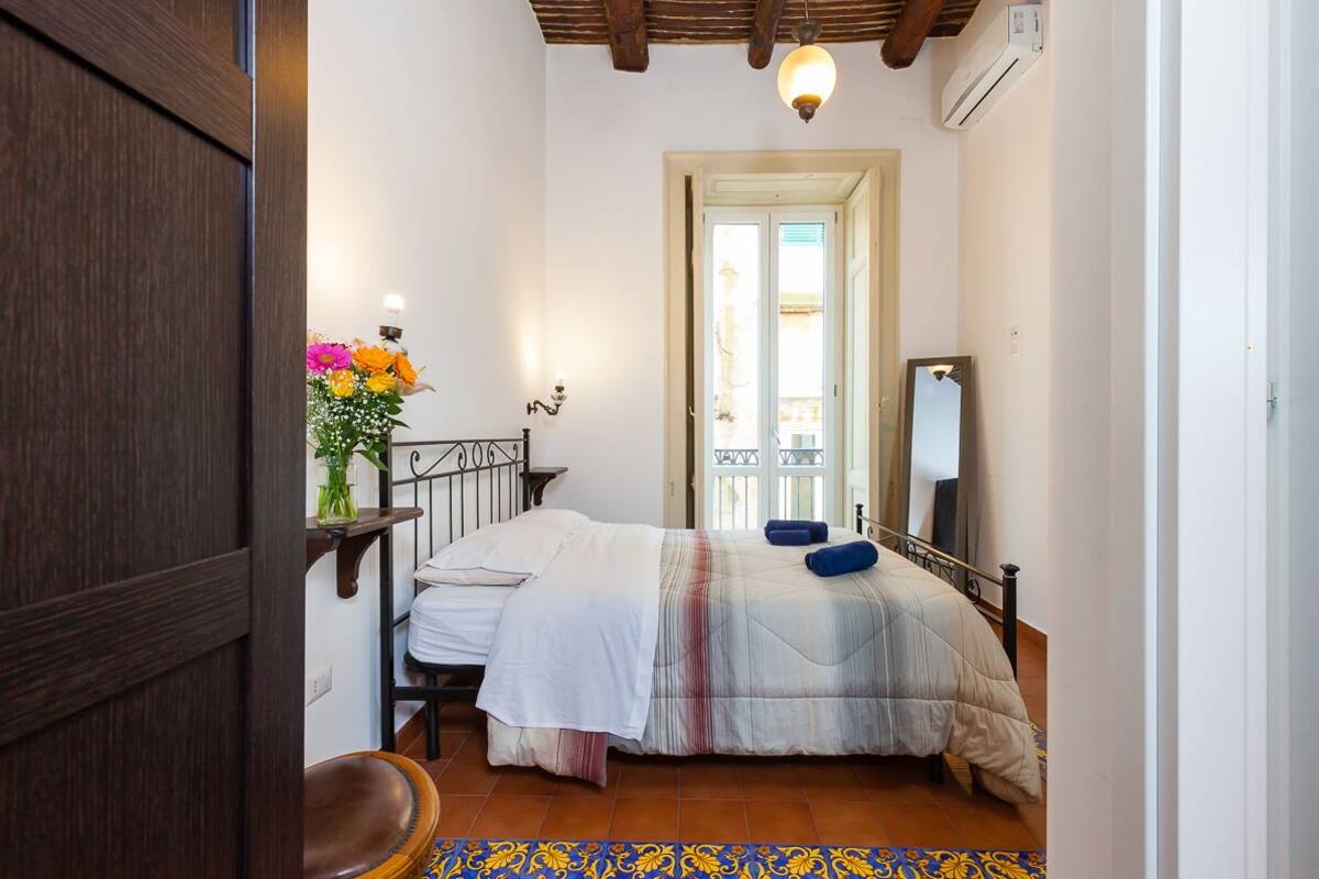 A bedroom with a bed, a wooden chair, a small table with a vase of flowers, a mirror, and a door leading to another room.