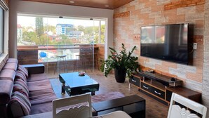 Una sala moderna con un sofá, una mesa de centro, un televisor montado en una pared de ladrillo y un balcón con vistas a edificios y árboles.