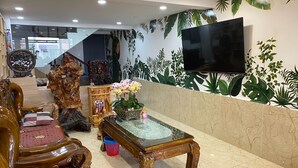 Una sala de estar con un sofá de madera, una mesa de centro de vidrio, un televisor montado en la pared y una pared decorativa con motivos vegetales.
