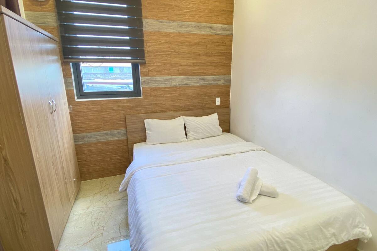 A small, neatly made room with a bed, a wooden wardrobe, and a window with blinds.