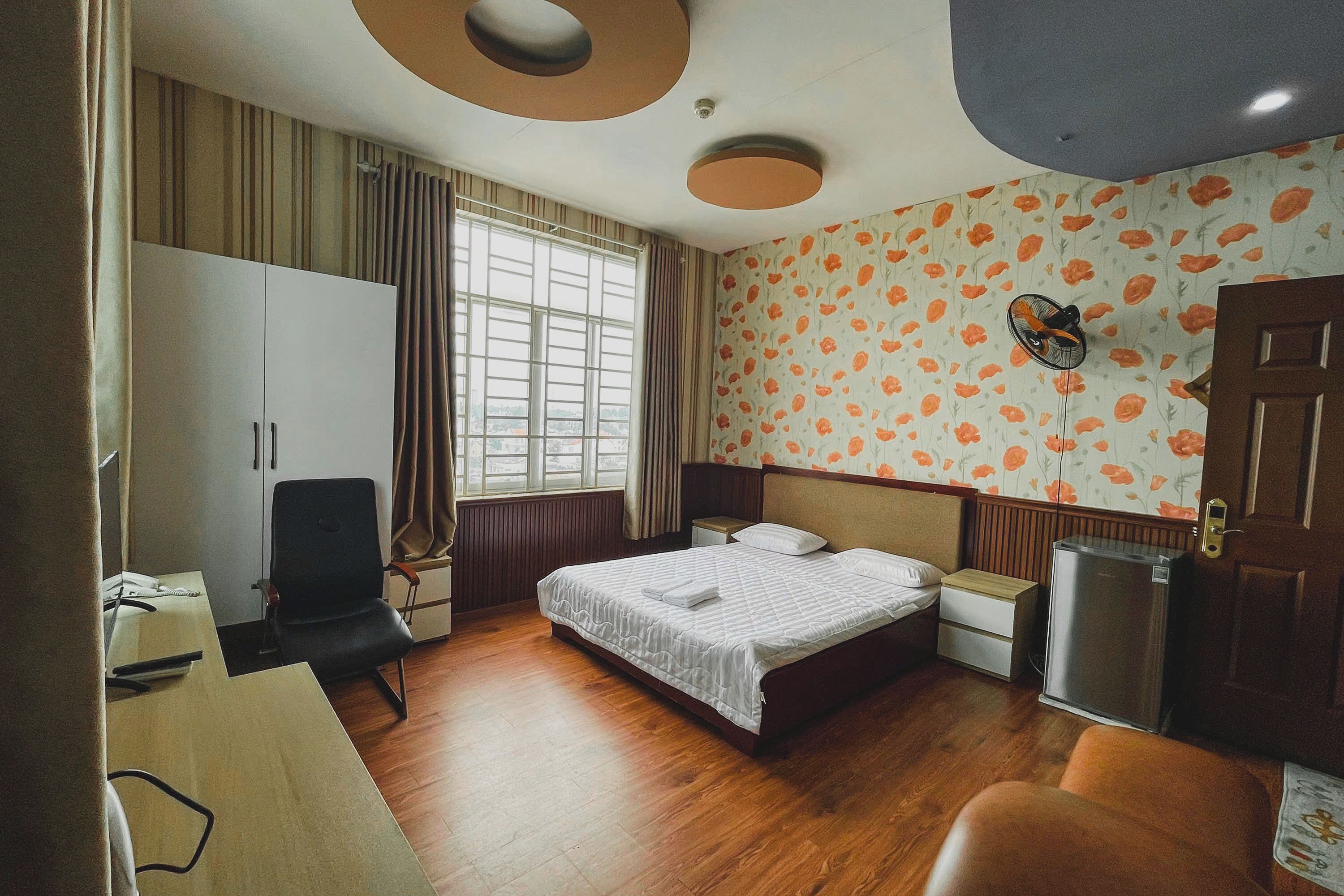 A hotel room with a bed, a desk, a chair, a wardrobe, a window with curtains, a wall with a floral pattern, and a ceiling fan.