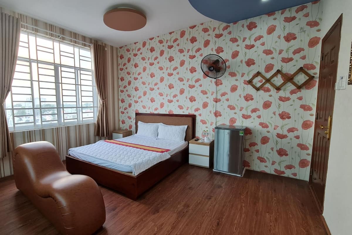 A hotel room with a bed, a chair, a bedside table, a refrigerator, a window with curtains, and floral wallpaper.