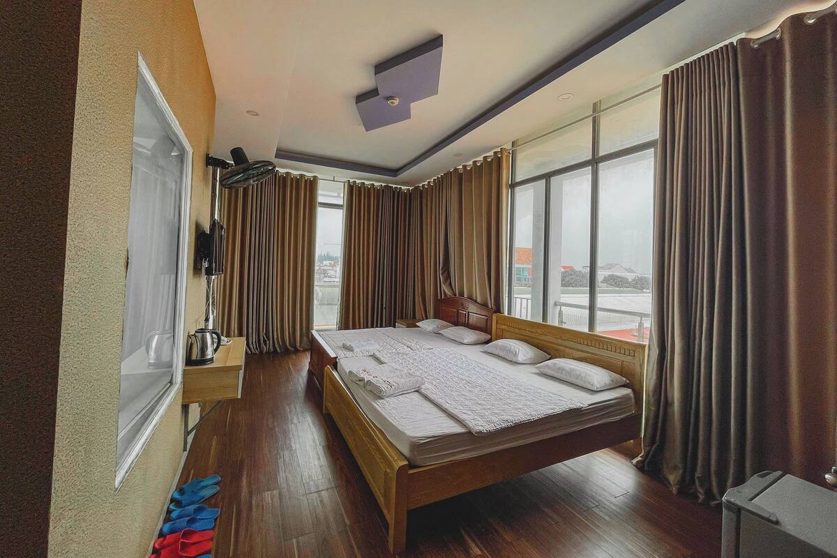 A hotel room with a large bed, a wooden headboard, a bedside table with a lamp, and a window with curtains.