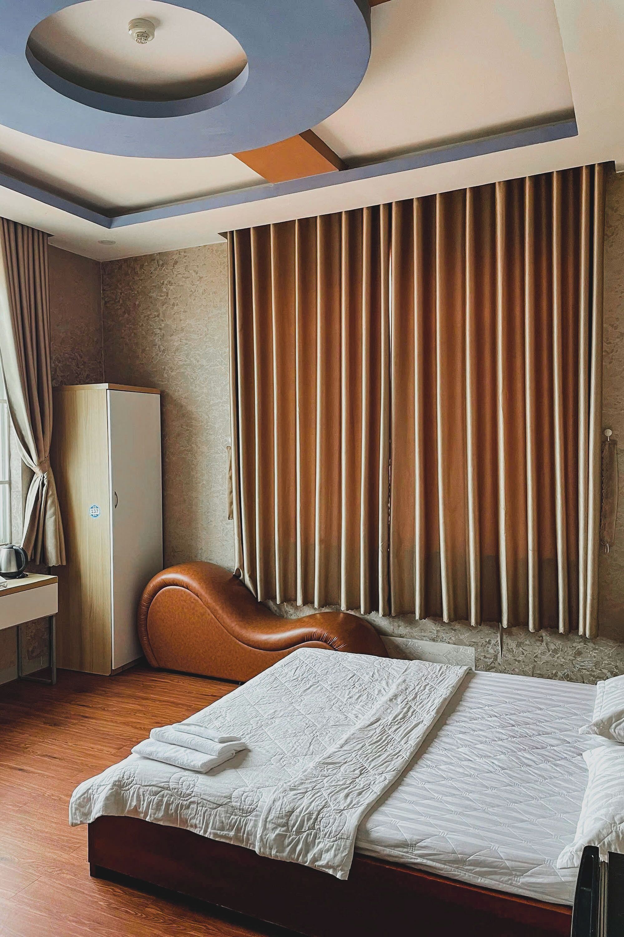A hotel room with a bed, a wardrobe, a brown leather chair, and a large window with curtains.