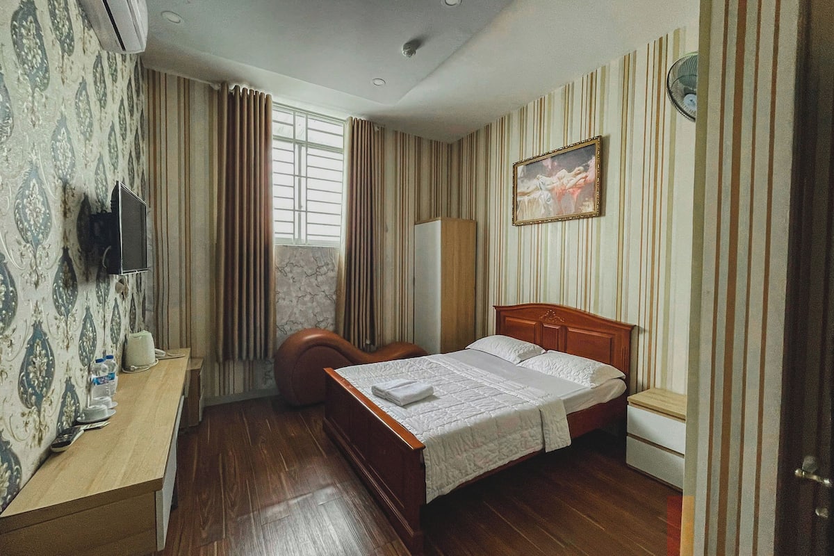 A hotel room with a wooden bed, a desk with a TV, a wardrobe, and a painting on the wall.