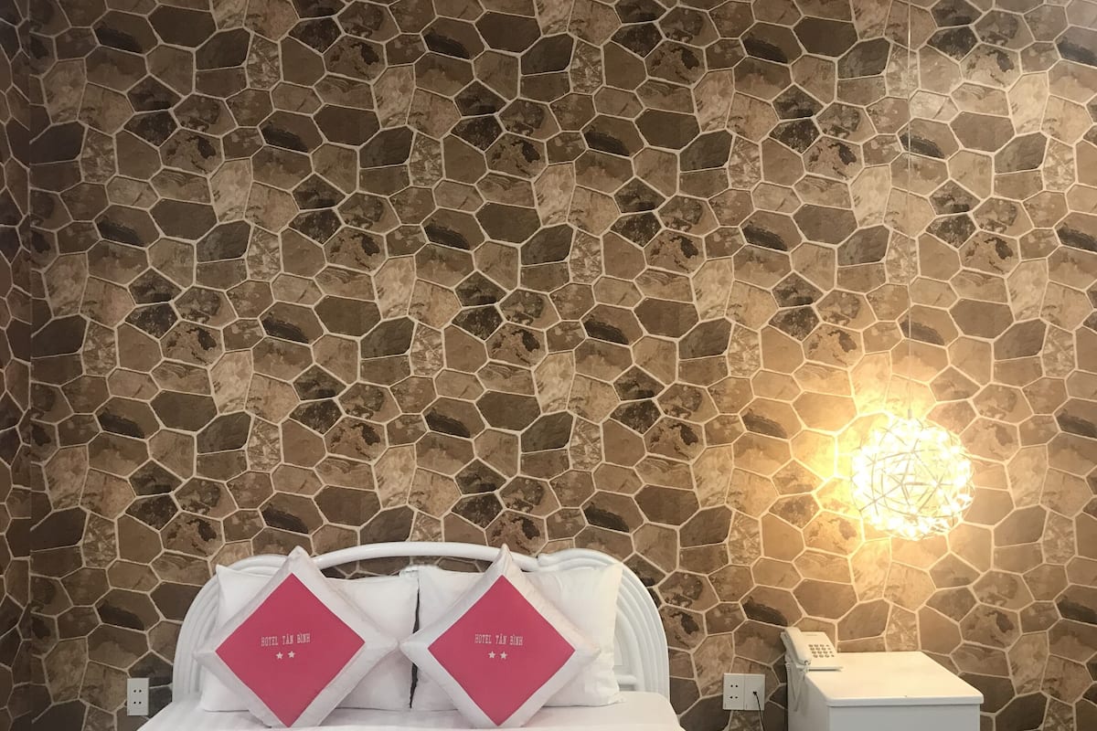 Een hotelkamer met een wand met een stenen patroon, een bed met witte lakens en roze kussens, en een nachtkastje met een telefoon.