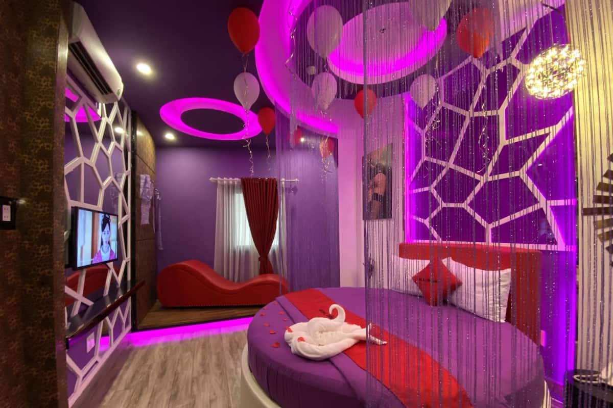 Een moderne slaapkamer met een gebogen bed, een televisie en decoratieve elementen zoals ballonnen en verlichting.