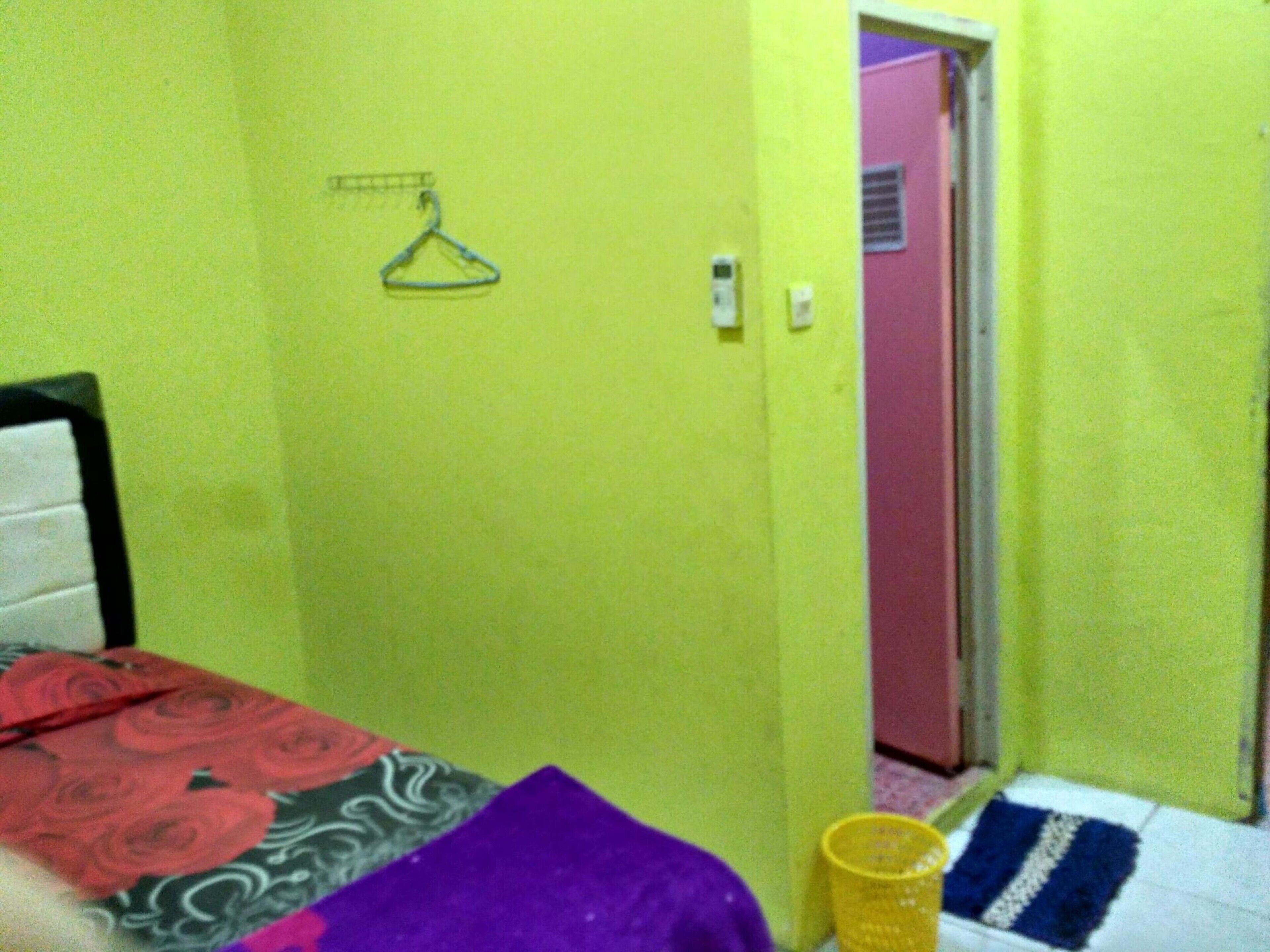 Una habitación pequeña y colorida con una cama, un bote de basura amarillo y una puerta que da a otra habitación.